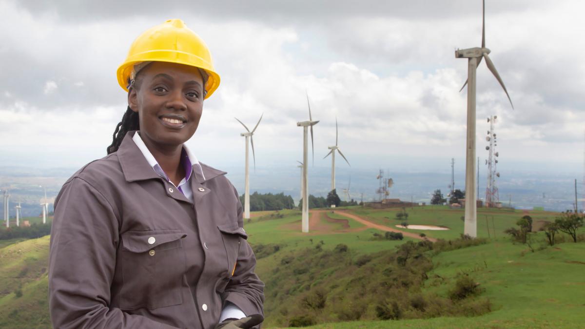 A smiling woman engineer in the foreground of a hilltop with several wind turbines