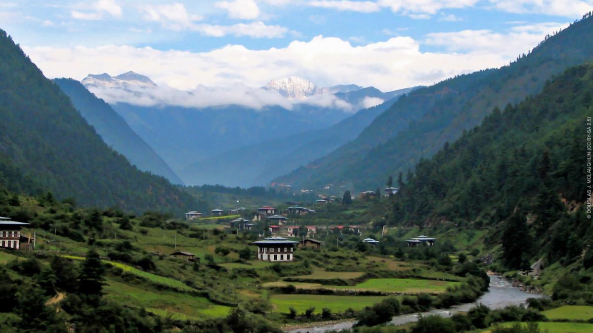 A village nestled in a green valley between high wooded mountains with the Himalayas in the distant backgorund
