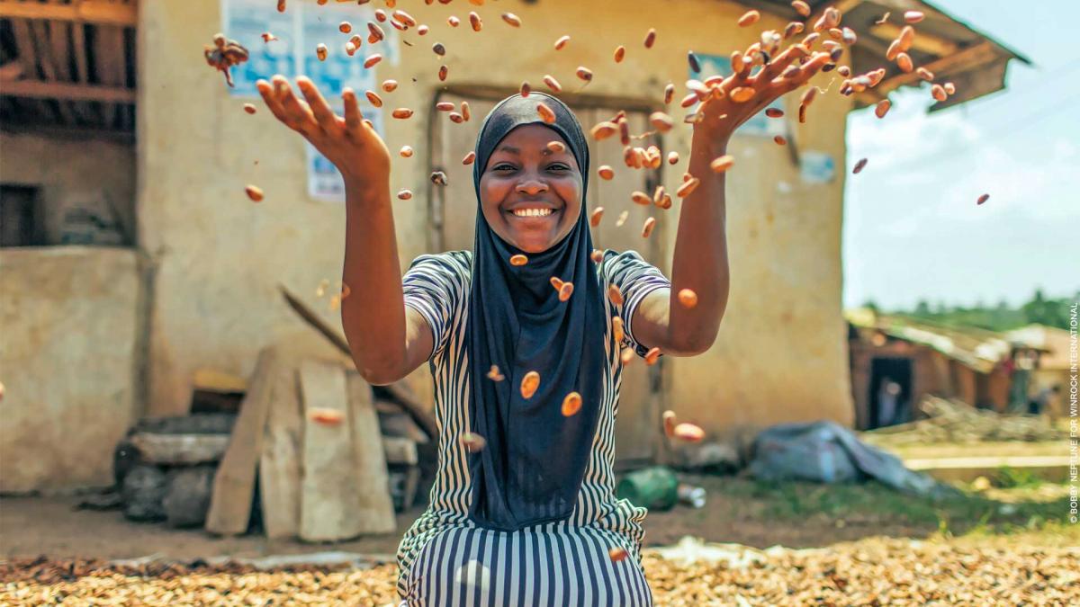 A smiling Nigerian woman tosses dried cocoa beans in the air 