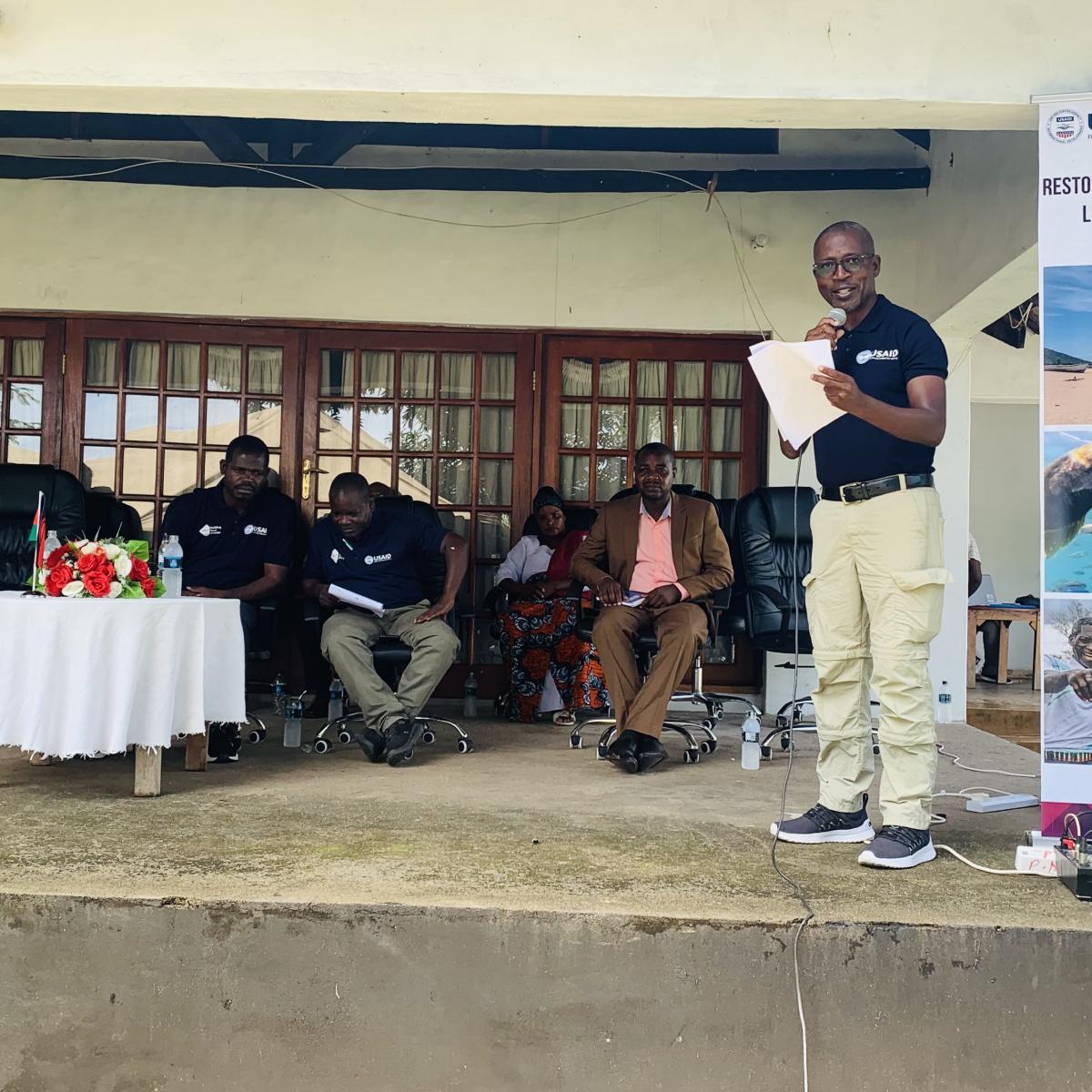 USAID Malawi Climate Change Specialist Bruce Sosola standing on an elevated area and giving remarks during the handover ceremony of an electronic catch assessment survey (eCAS) system and refurbished boats to the Government of Malawi.