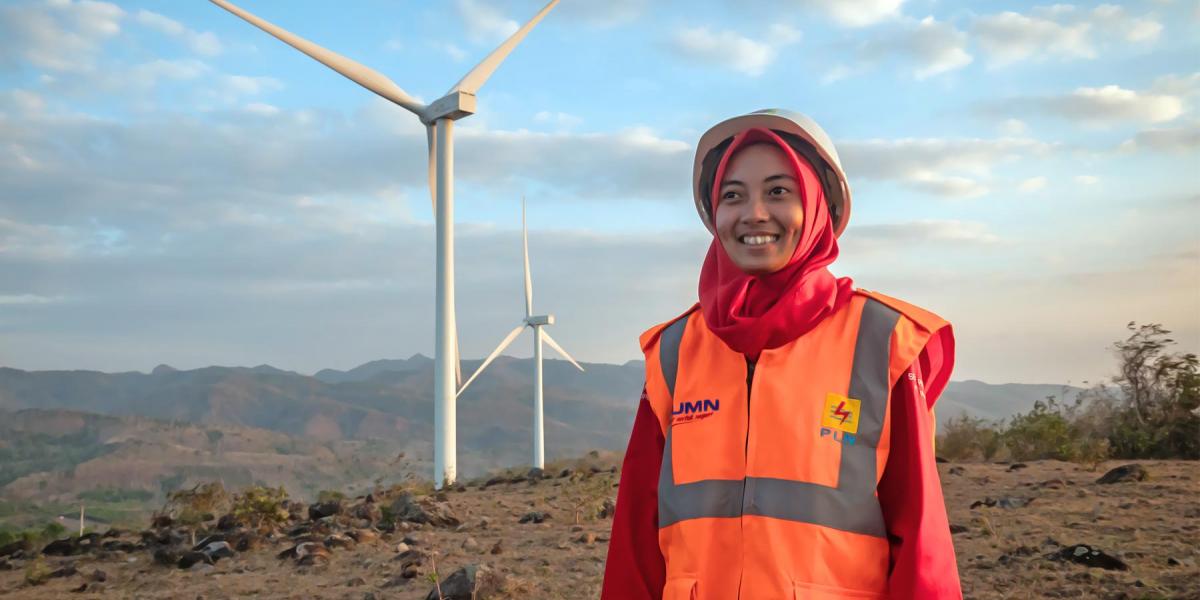 A smiling female renewable energy engineer stands in front of the large turbines on a wind farm.
