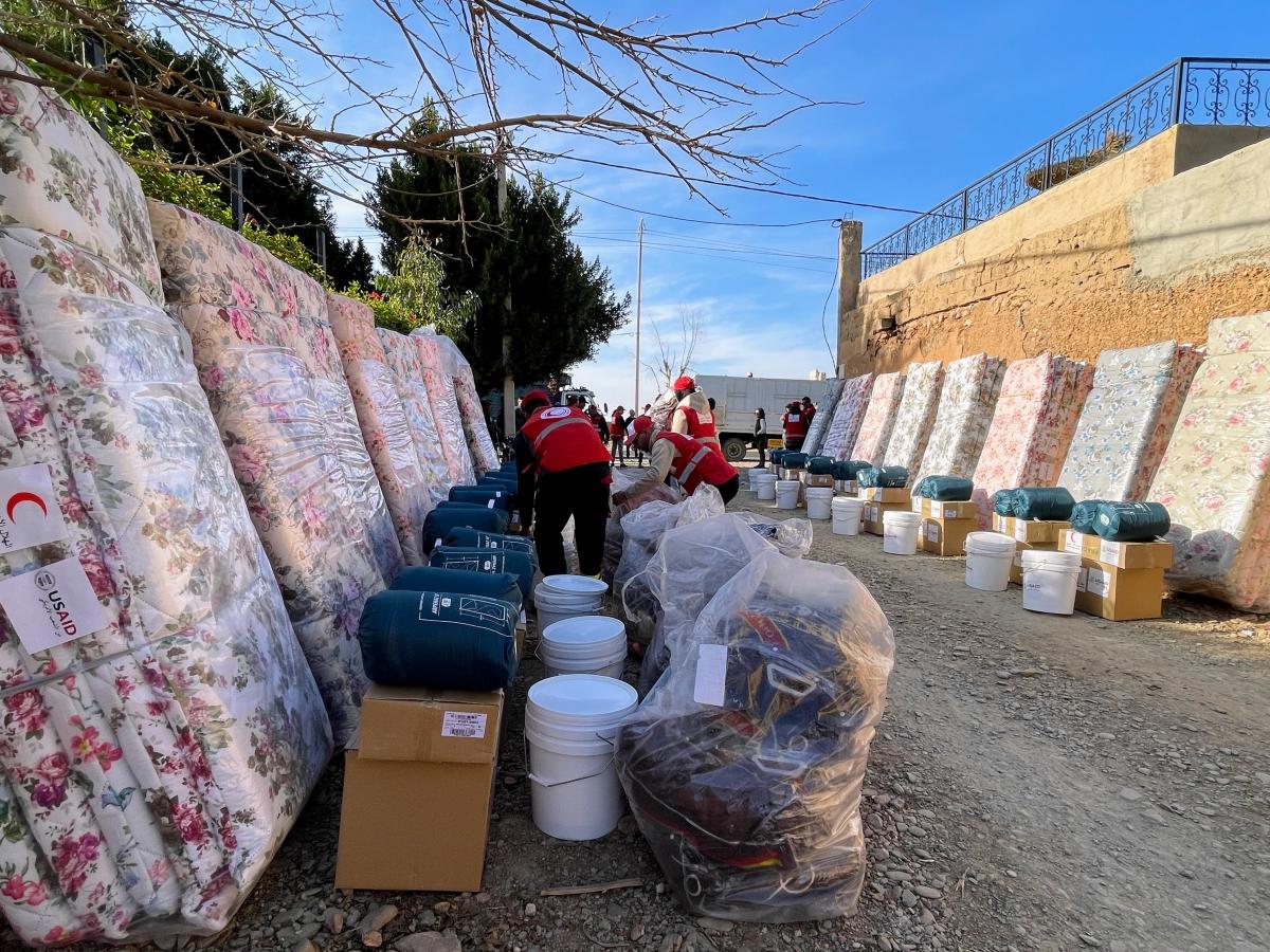 Packages of household essentials, including mattresses, bags of blankets, boxes, and buckets, are lined up along exterior walls on both sides of a gravel road.