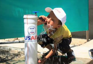 A young boy in a USAID baseball cap drinks directly out of a water pump