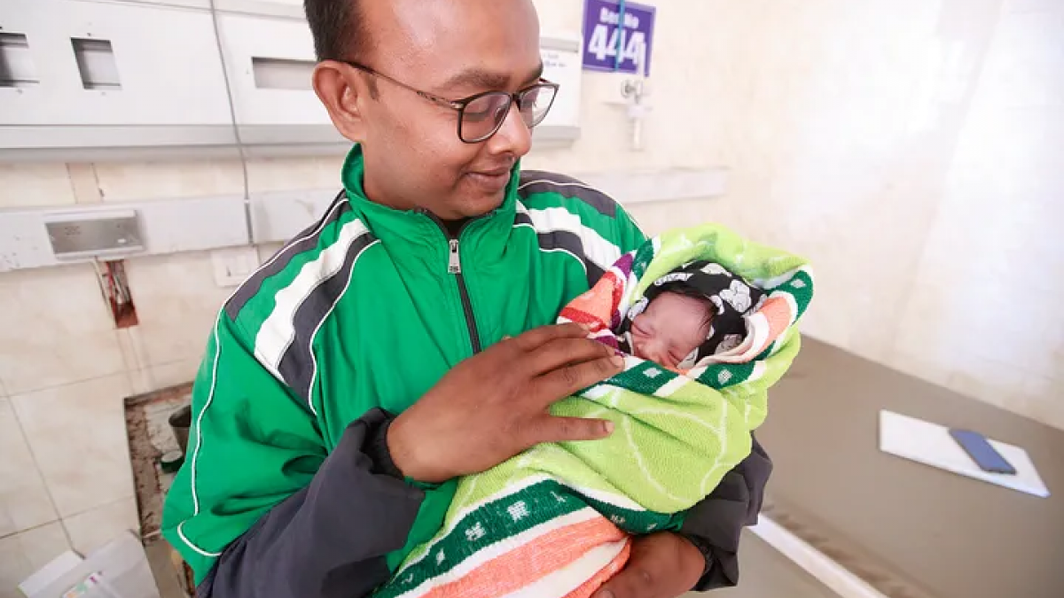 A man, wearing a green athletic jacket and glasses holds, his newborn who is swaddled in a blanket. 