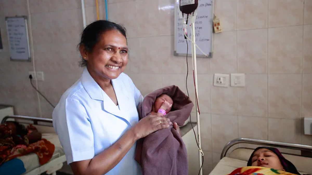 Health worker wearing a nun's suit dress holds a baby while smiling over a mother sleeping a cot. 