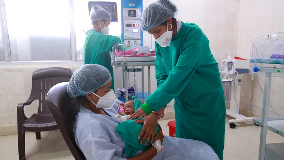 Three people wearing personal protective equipment in a hospital treatment room. Two health care workers help woman holding anewborn.