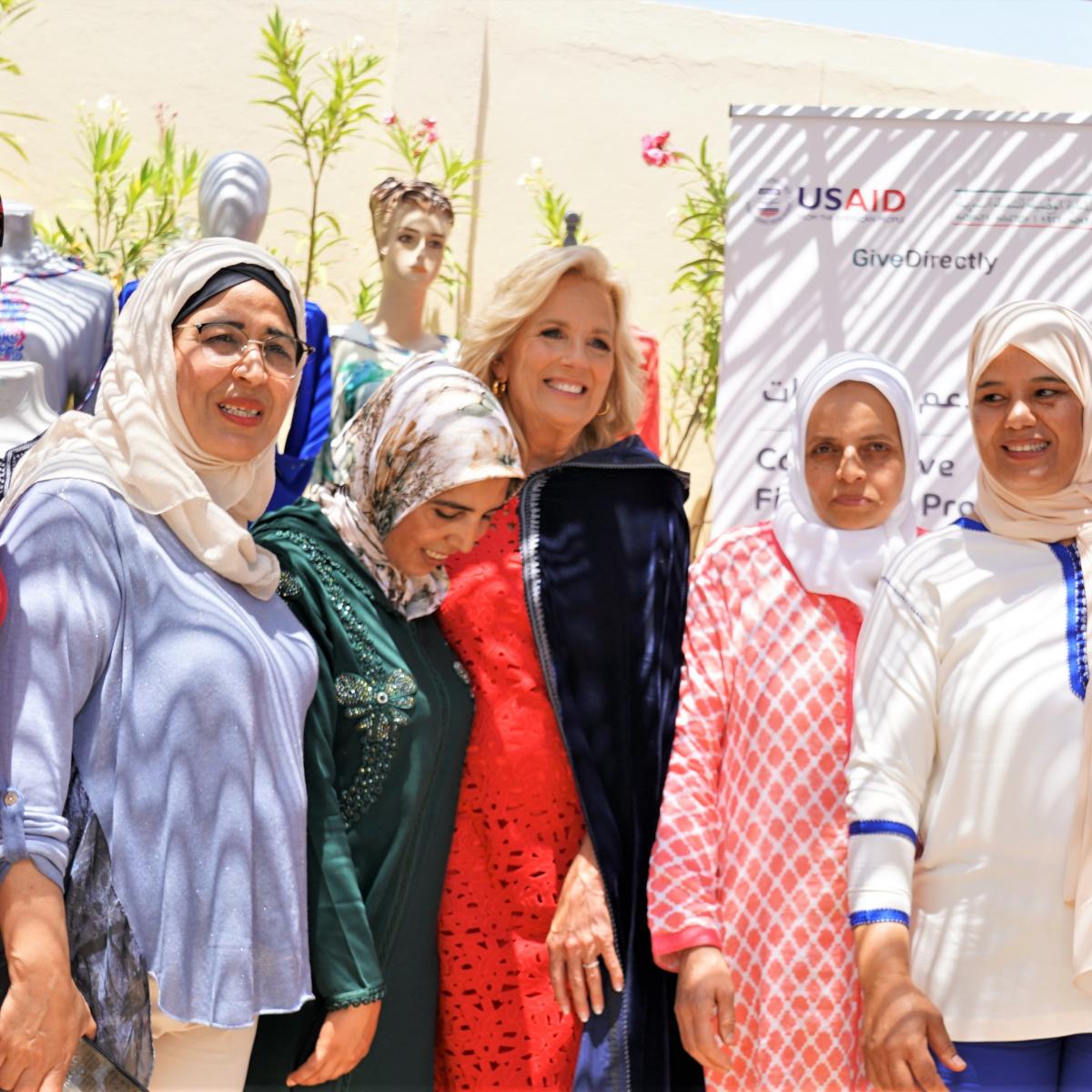 A group of women, mostly hijab-wearing, pose with First Lady Dr. Jill Biden, who is wearing a traditional Moroccan cape that is blue. A USAID project banner is displayed behind them, and it is sunny.