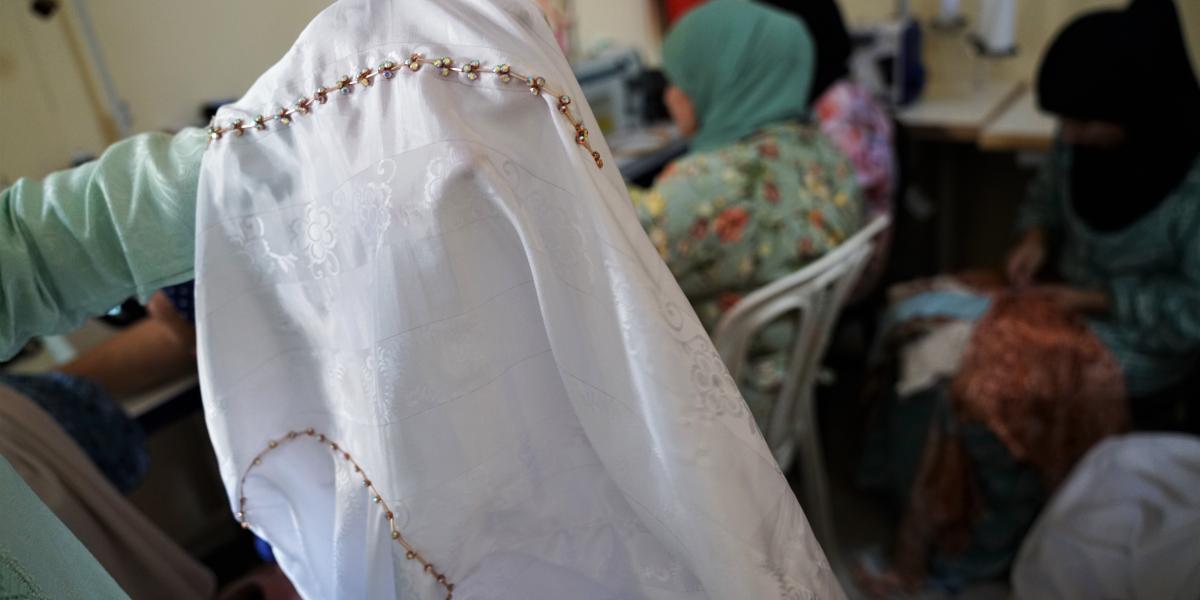 A white traditional Moroccan dress is held up to demonstrate the details of its jeweled trimming. Women wearing hijabs work in the background, facing away from the camera.