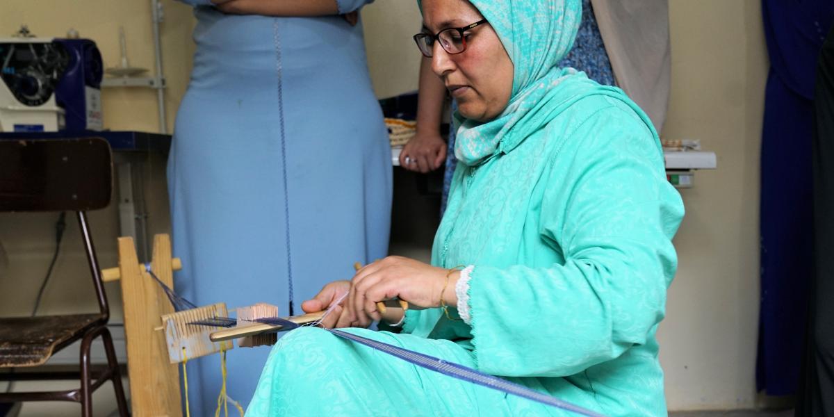 A Moroccan woman in aqua-colored dress and hijab sits and works with a specialized embroidery tool.