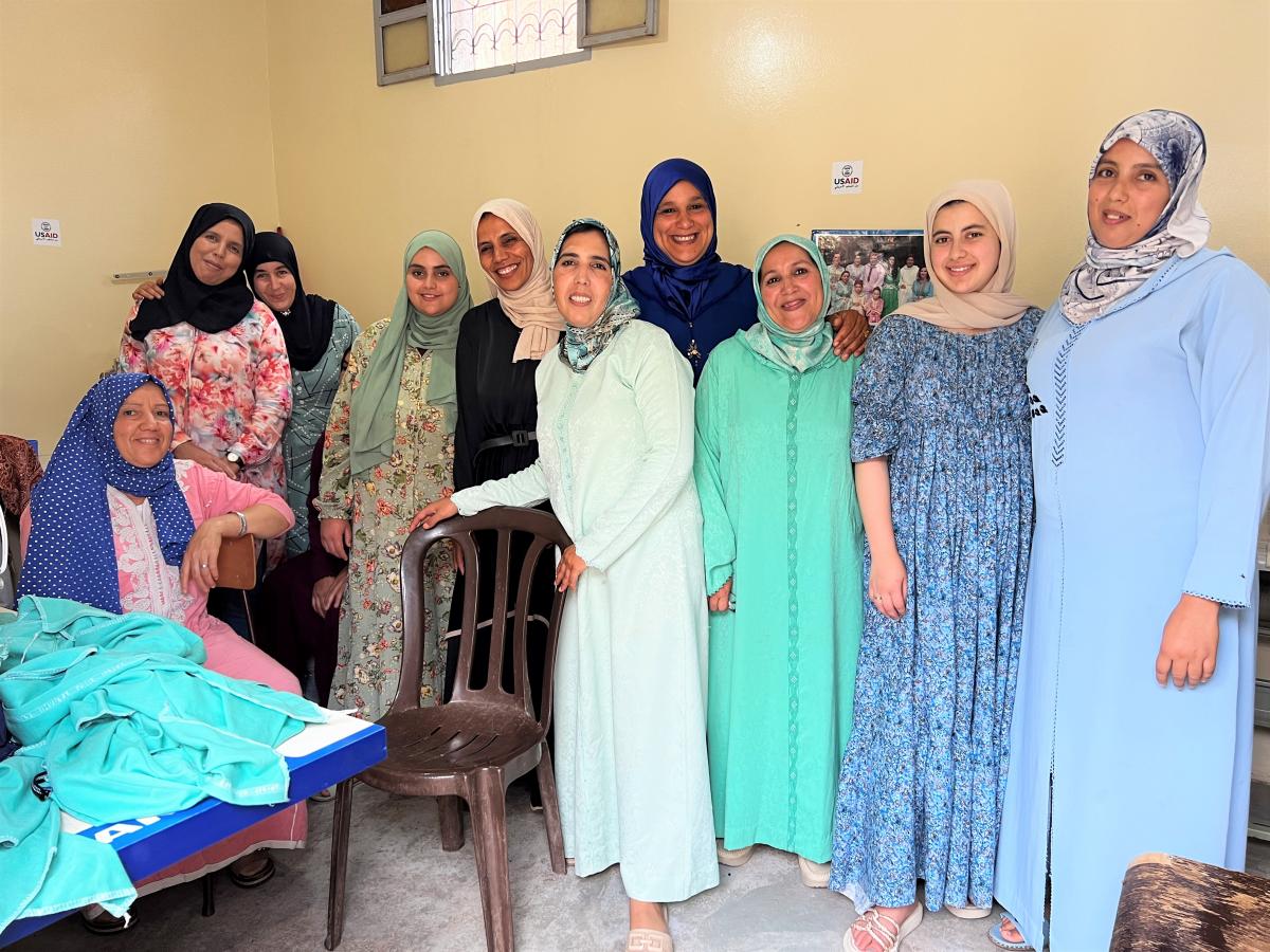 Ten Moroccan women in colorful dress, all wearing hijab, pose in their workshop facing the camera.