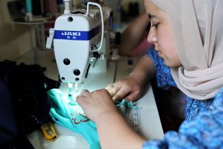 A young woman wearing a blue dress and beige hijab stiches an item of clothing with a sewing machine.