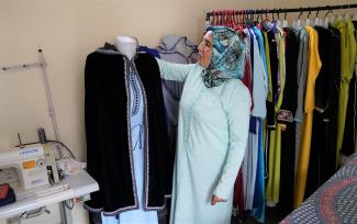 A Moroccan woman in a light blue dress and multi-colored hijab inspects a traditional Moroccan cape on a mannequin. A sewing machine can be seen in the lower left corner, and a variety of traditional dresses hang on a rack behind the woman. 