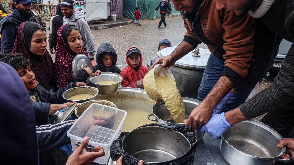 Two men pour lentils into a pot from a large vat while children circle them with their containers outreached waiting for their share.