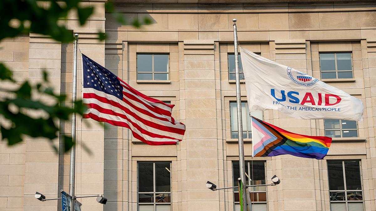 A Progress Pride flag flying alongside American and USAID flags in front of an office building.