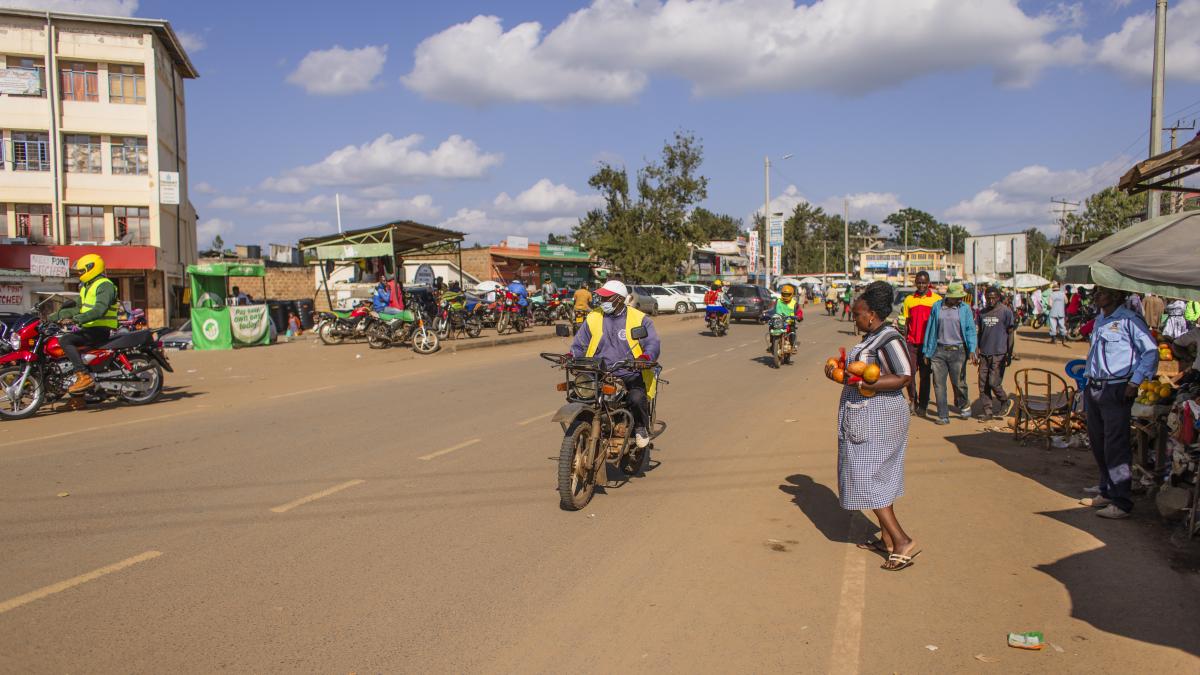 A motorcycle pulls up to a woman holding fruit she has bagged ahead for the driver.