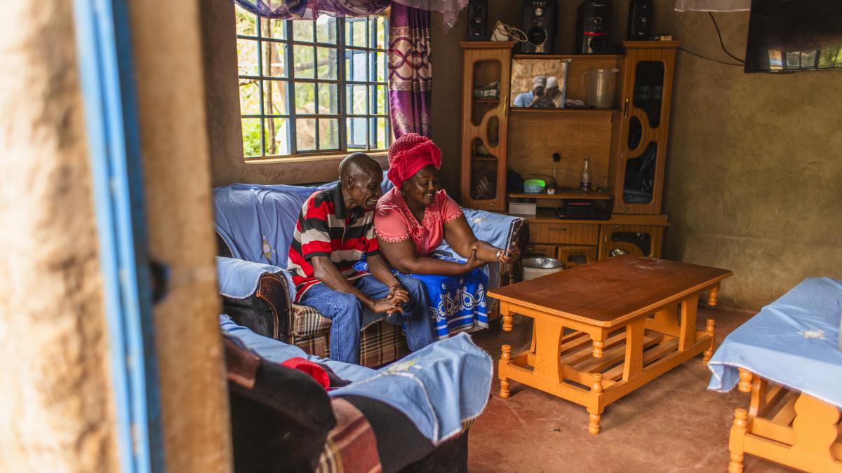 A man and woman sit smiling in their home while talking with their son via smartphone.