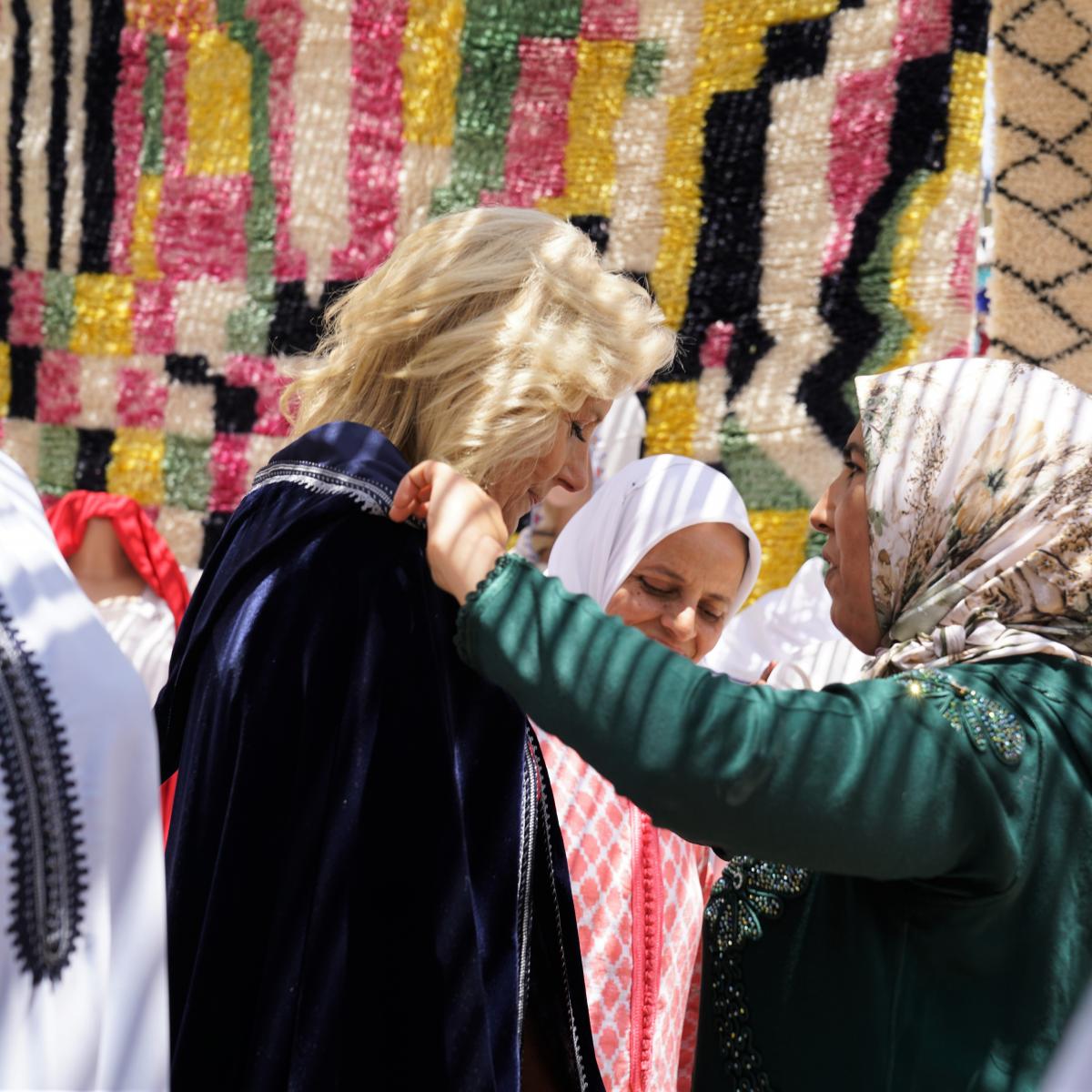 A woman in green traditional Moroccan dress and a beige hijab, right, fastens a traditional blue cape over the shoulders of U.S. First Lady Dr. Jill Biden, left, in front of Moroccan rugs that hang in the background.