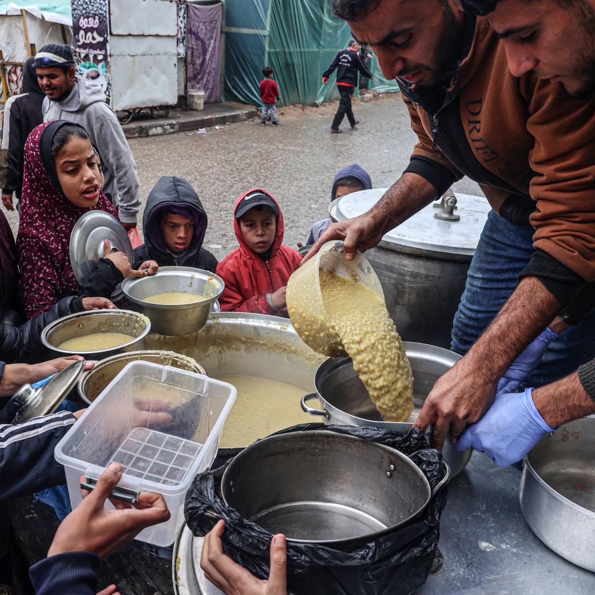 Two men pour lentils into a pot from a large vat while children circle them with their containers outreached waiting for their share.