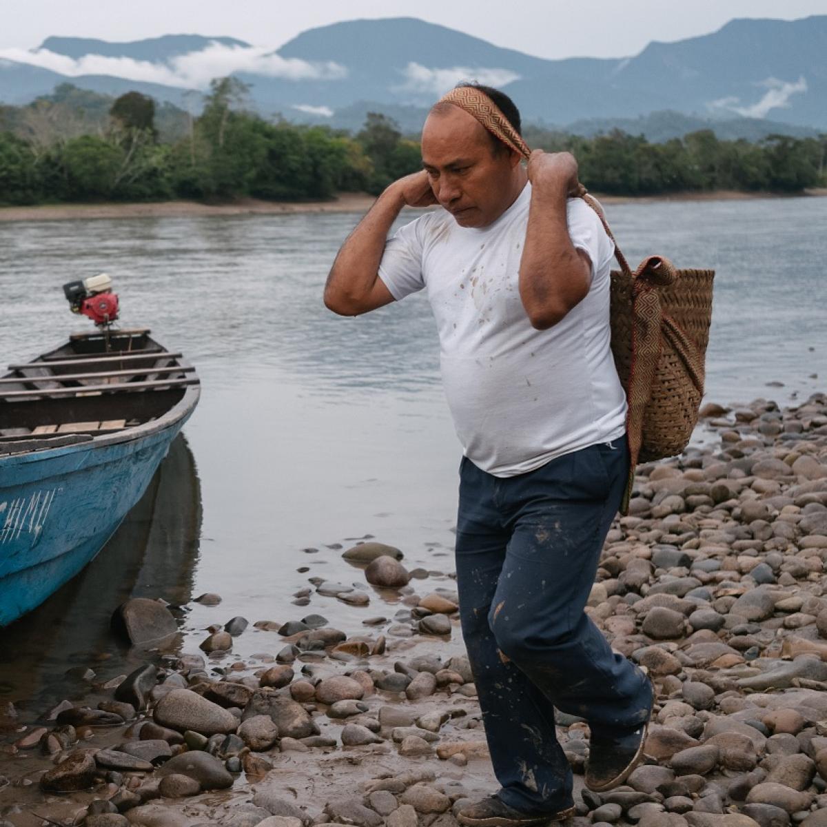 A man walks along a river shoreline carry a sack slung across his forehead.