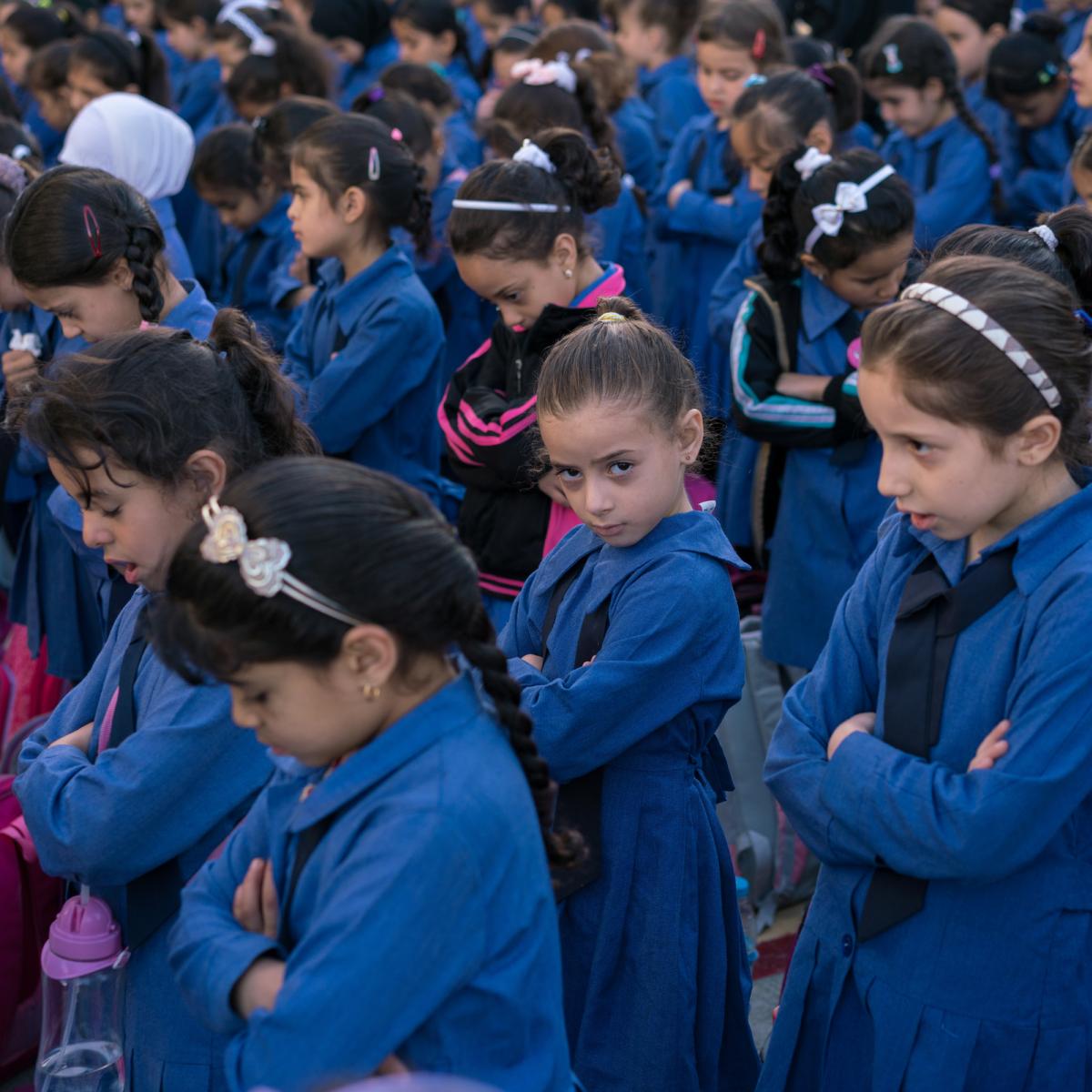 A girls looks up among a crowd of girls with their heads bowed.