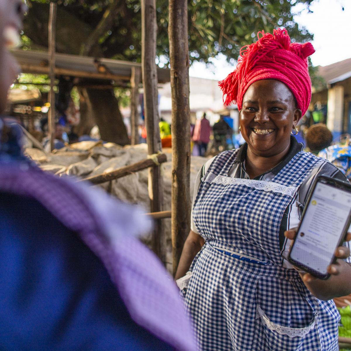 A woman holds a smartphone up so a vendor can see the screen while at a market in Kenya.