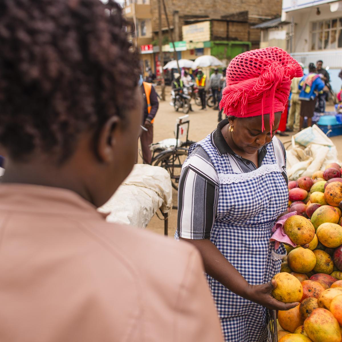 A woman with her back to the camera inspects fruit while another woman prepares her order.