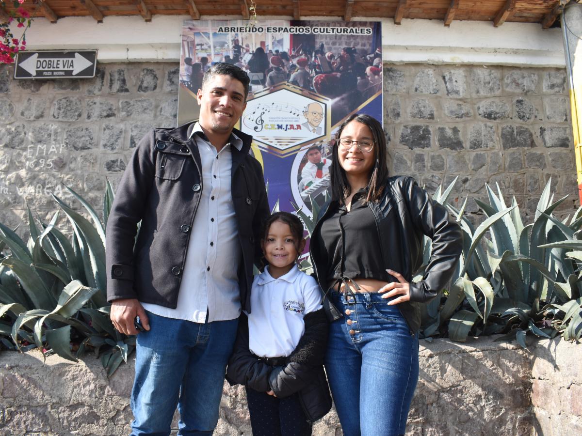 This picture shows Daniel Bracho, his wife and daughter, all three standing in front of the music school sign. The background shows a stone wall with agave plants planted. 