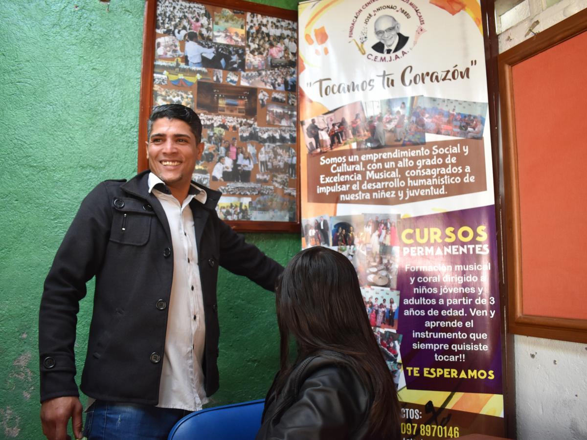 This picture shows Daniel Bracho in a black coat, proudly showing a roll-up banner with pictures and information on the school. He is standing up, smiling. His wife, Mirvic, is sitting down, close to him looking at the banner. 