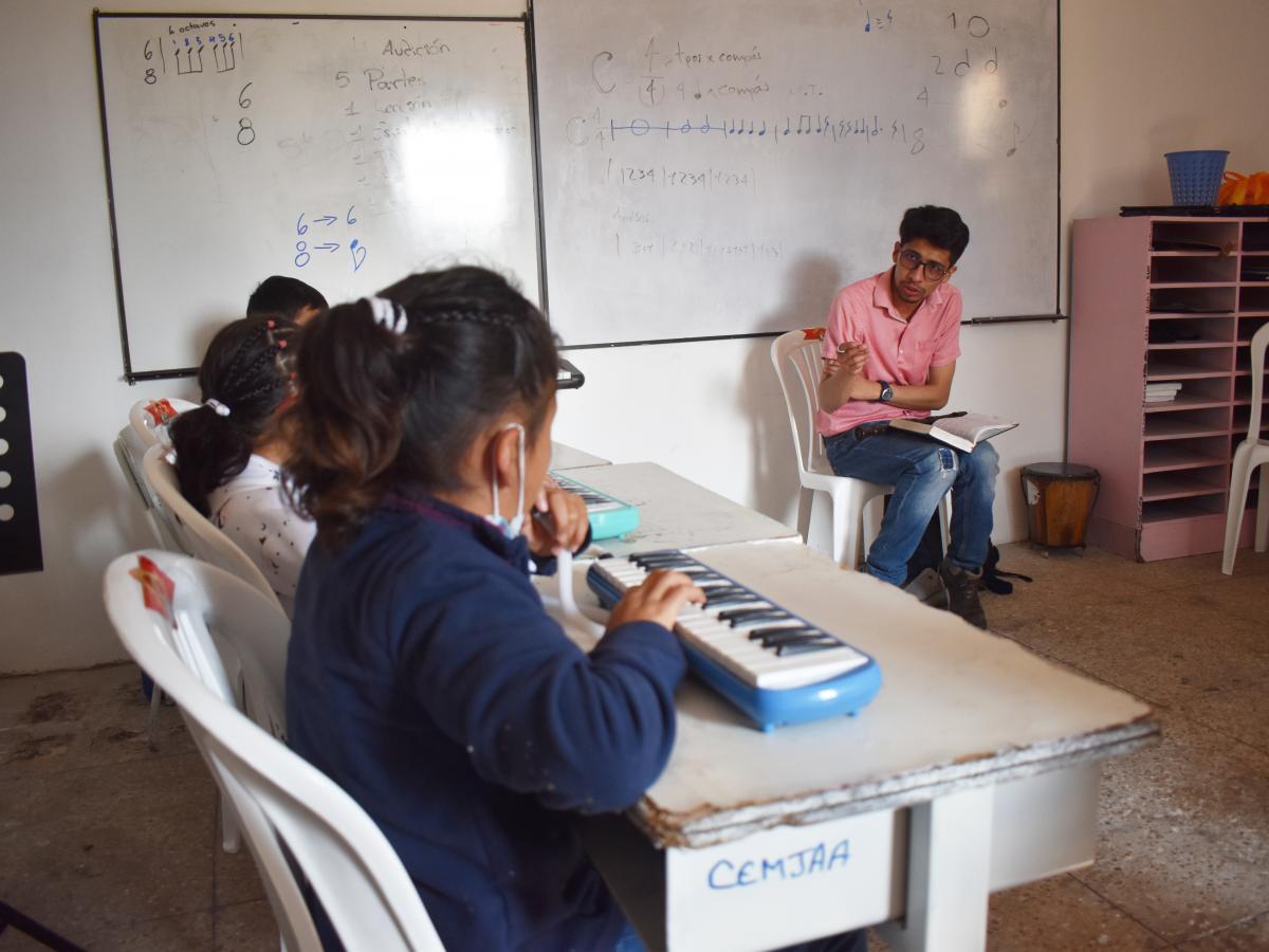 This picture shows the back of three children while learning to play the piano. The teacher is in front of them, in the back of the room instructing them to do something. 