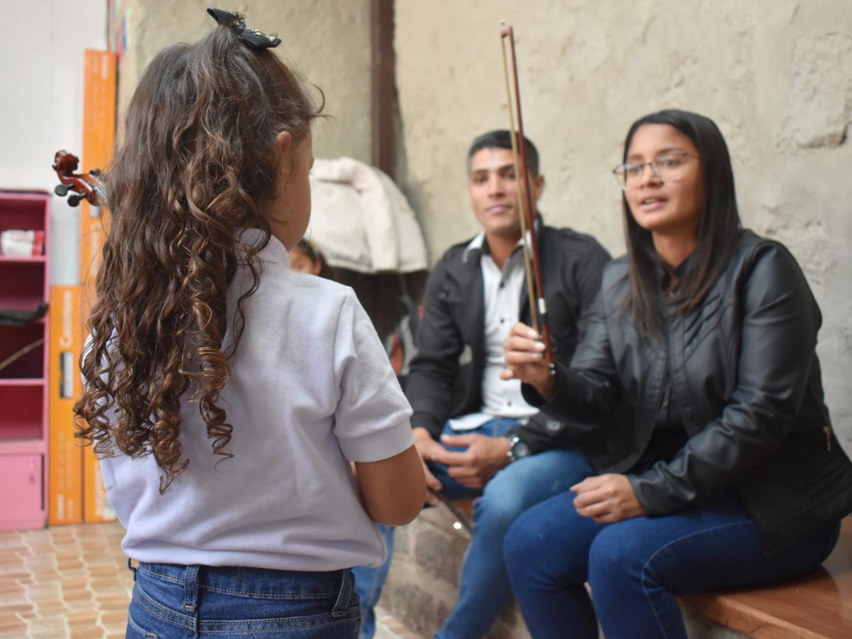 This picture shows Daniel Bracho and his wife, Mirvic, teaching a little girl with curly hair to play the violin. Mirvic is holding the bow and looking attentively at the girl. 