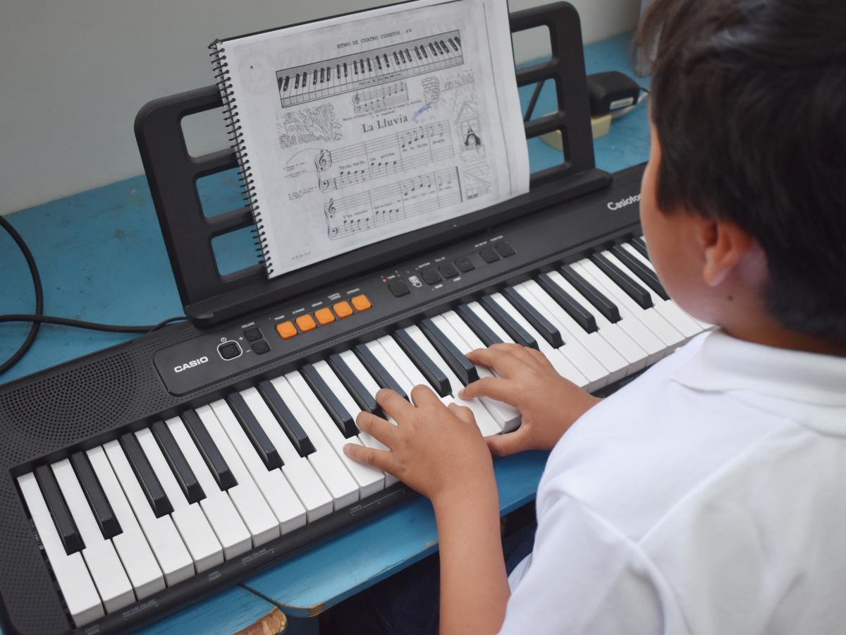 This picture shows the back of a child with short hair playing the piano. There is a booklet with notes on a scale that he is following. 
