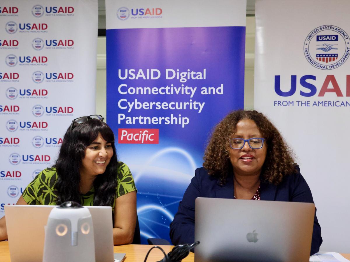 Left to right is USAID DCCP-Pacific Chief of Party Anju Mangal and USAID Pacific Islands Mission Director Zema Semunegus sitting on a desk where Semunegus delivered opening remarks at the USAID DCCP-Pacific and the Asia Open RAN Academy virtual partnership signing ceremony. USAID pull up banners are placed behind the women’s seat.