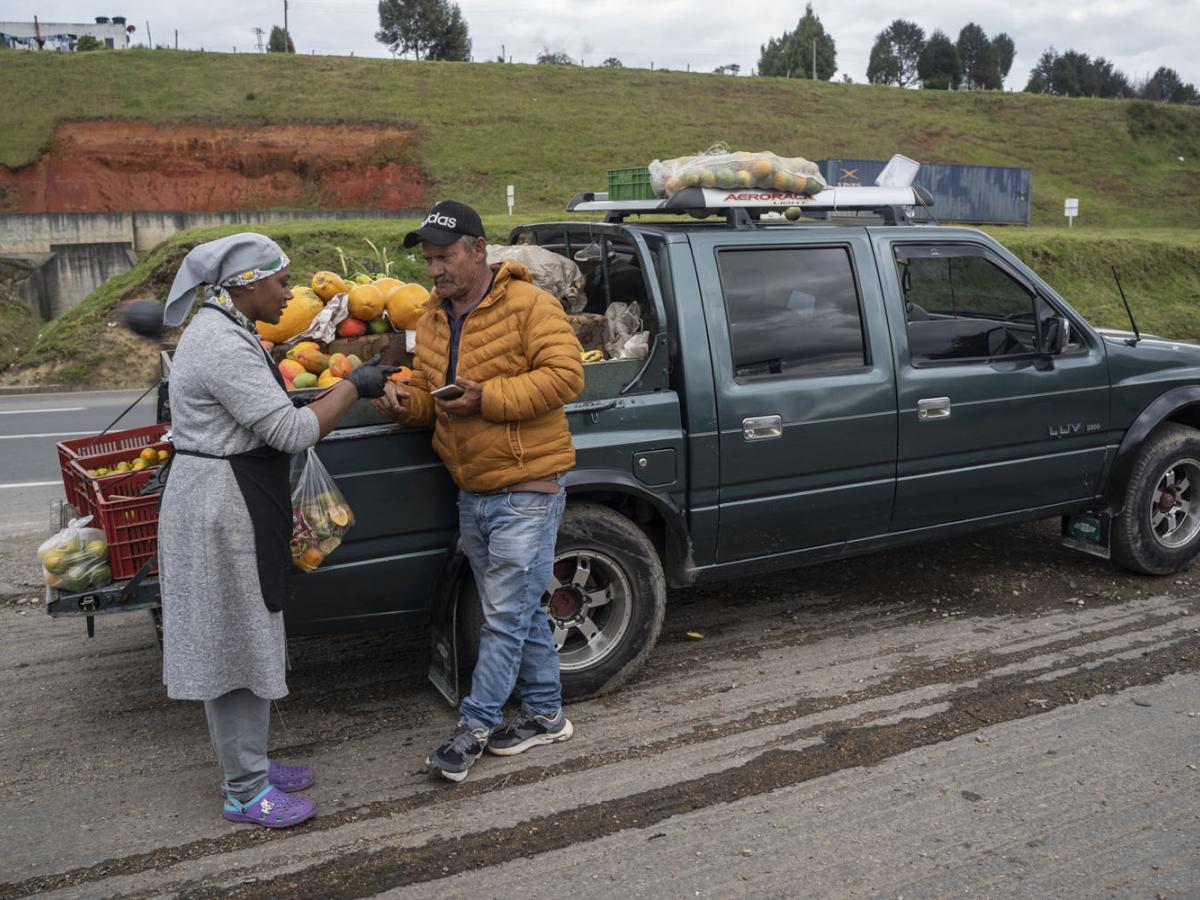 Angie stands next to a pickup truck holding a bag of produce she is purchasing from the local vendor in front of her. The vendor uses his phone to complete the transaction.