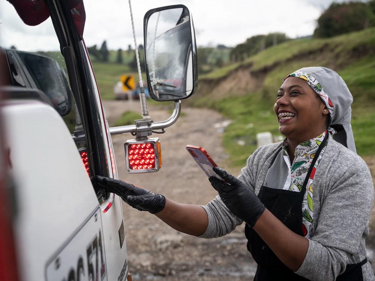 A smiling Angie stands in her chef uniform looking through a truck window while holding her cell phone in her hand. 