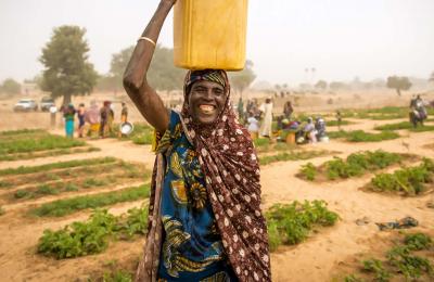 A smiling Nigerian woman carries a bucket of water in a community garden