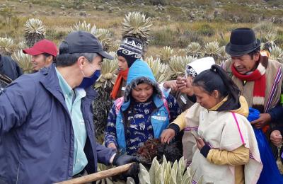 Two indigenous Guambiano women inspect soil carbon with a USAID scientist in a natural mountainous landscape. A number of other indigenous people and USAID technicians surround them.