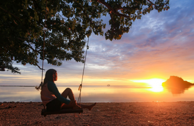 A young woman sits on a swing overlooking the setting sun over the water.