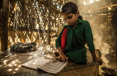 A young girl reads a book on a mat in a hut.