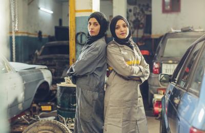 Two female auto shop workers wearing hijabs stand back-to-back in a shop garage.