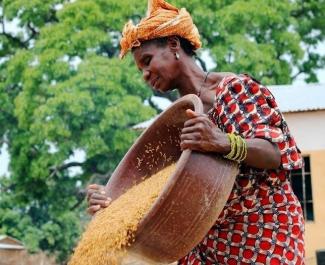 A Malian woman in a red patterned dress and orange headband emptying rice our of a large clay bowl. 