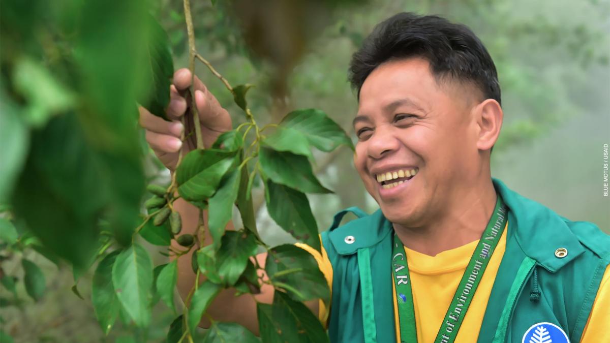 A smiling man in a forest patroller uniform examines a tree.