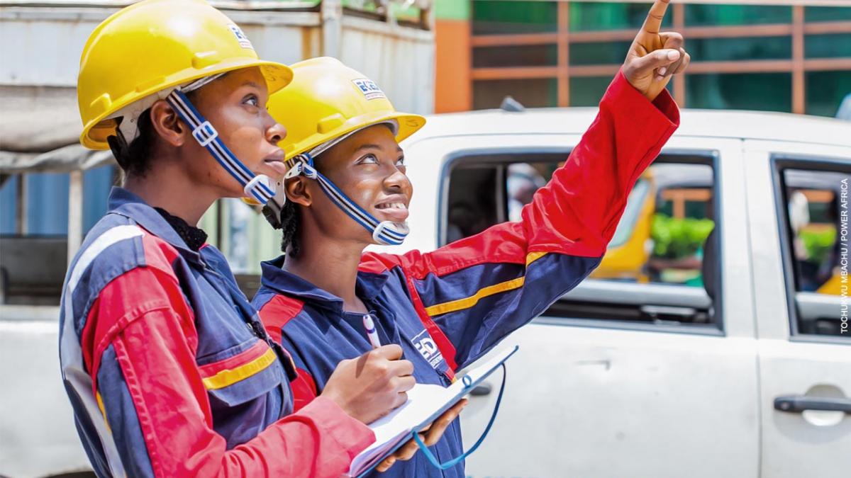 Two female electrical engineers dressed in power company safety gear inspect a power installation from the ground.