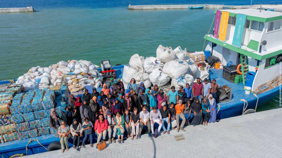 a large group of people pose at the edge of a boat full of bundled plastic waste in a tropical harbor