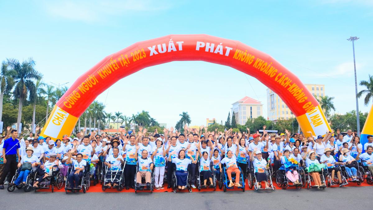 Hundreds of people, some of whom are wheelchair-users, excitedly prepare at the starting line of a race track. There is a large inflatable orange arch behind the racers contrasting with a bright blue sky and green palm trees.