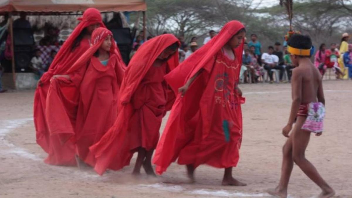 A procession of four women wearing red dresses and veils moving forward while a crowd watches.
