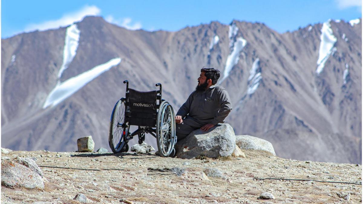 A man with black hair and a beard in a gray shirt and trousers sits on a large rock beside his wheelchair. In his background is a large jagged mountain range spotted with patches of snow and a bright blue sky. 