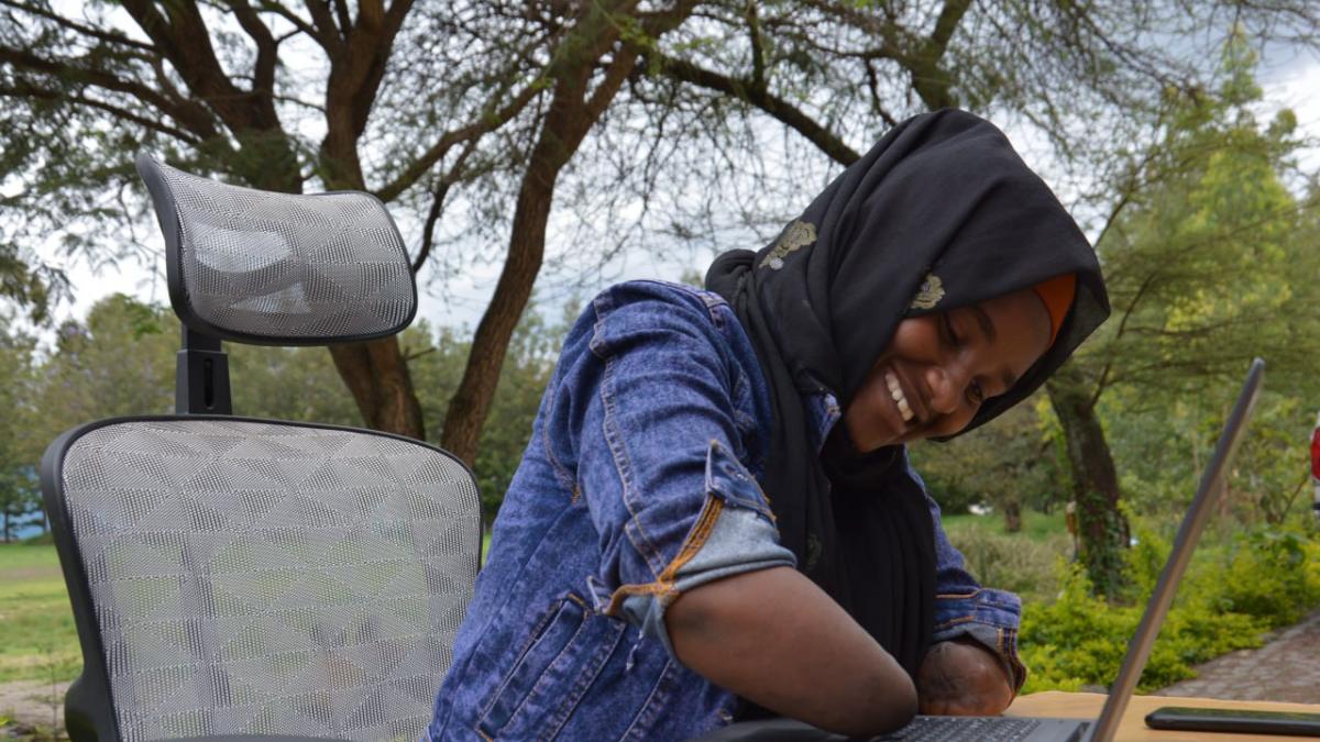 A young Ethiopian woman who is an amputee, wearing a black headscarf and denim top using a laptop outdoors smiles.
