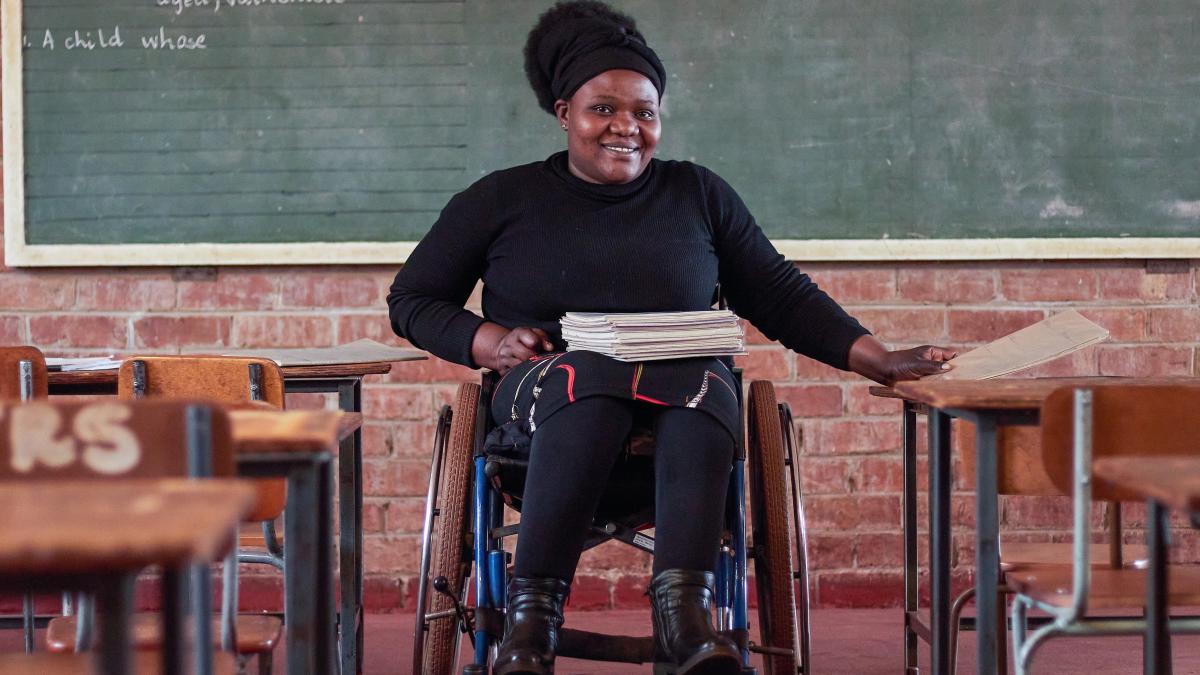 A Zimbabwean woman dressed in a black sweater, black tights, and a black, white, and red skirt with black boots sits in her wheelchair smiling at the camera. She has a pile of notebooks on her lap and is in front of a chalkboard in a classroom.