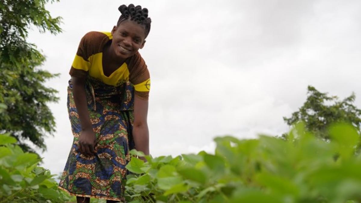 A young women smiles while kneeling down to display a field of green soybean plants