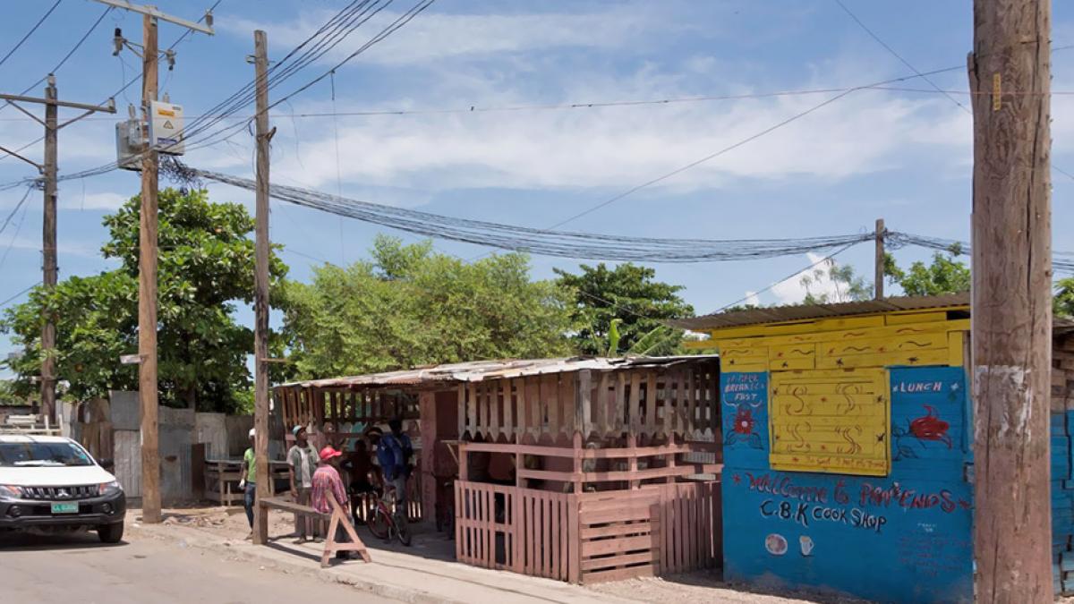 A small group of people in a poor village congregate casually outside a local shop under electricity distribution poles and a mass of associated wires.